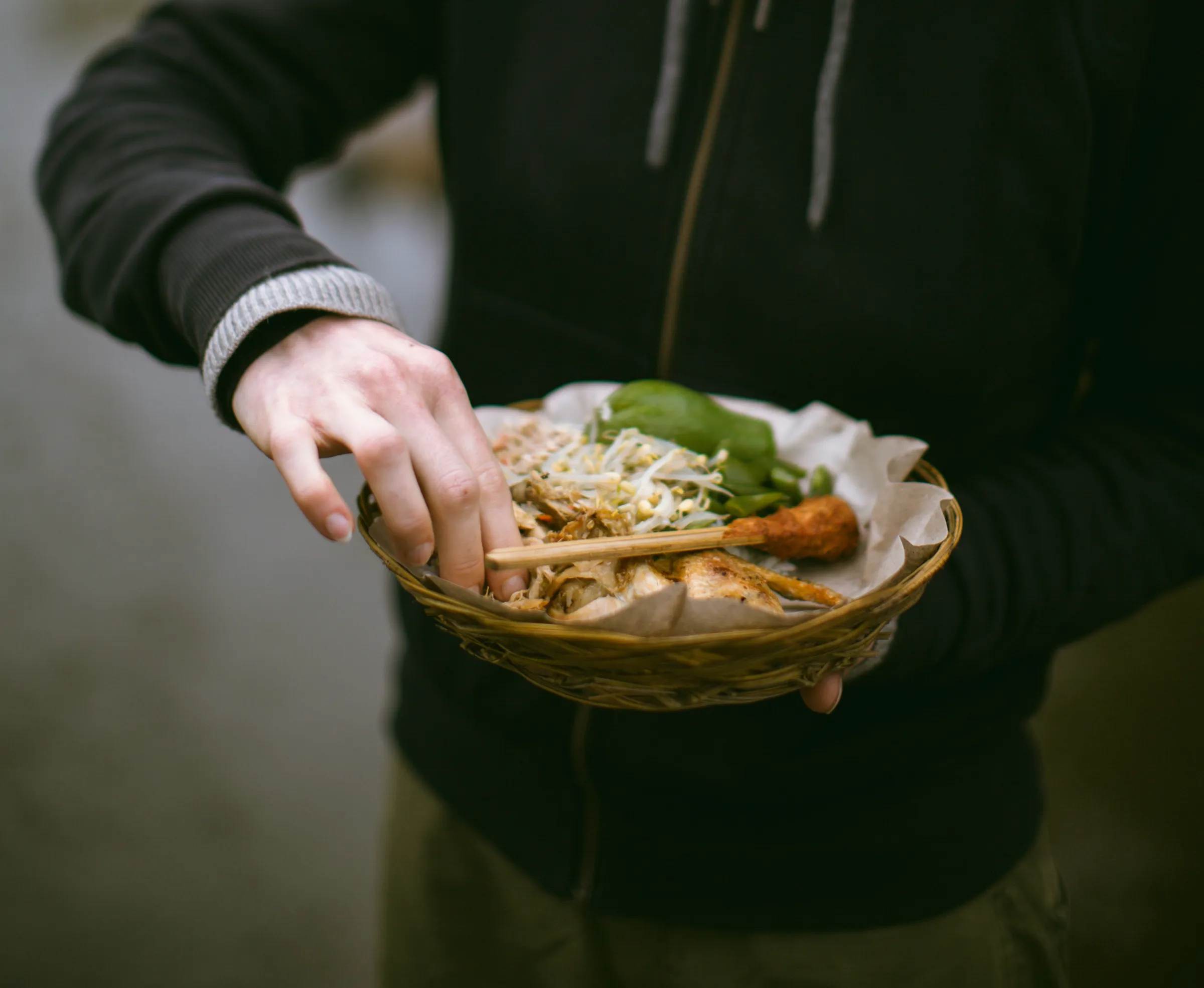 A woven basket of Balinese home cooking in a guest's hands: blanched vegetables, sprouts, sate and grilled chicken