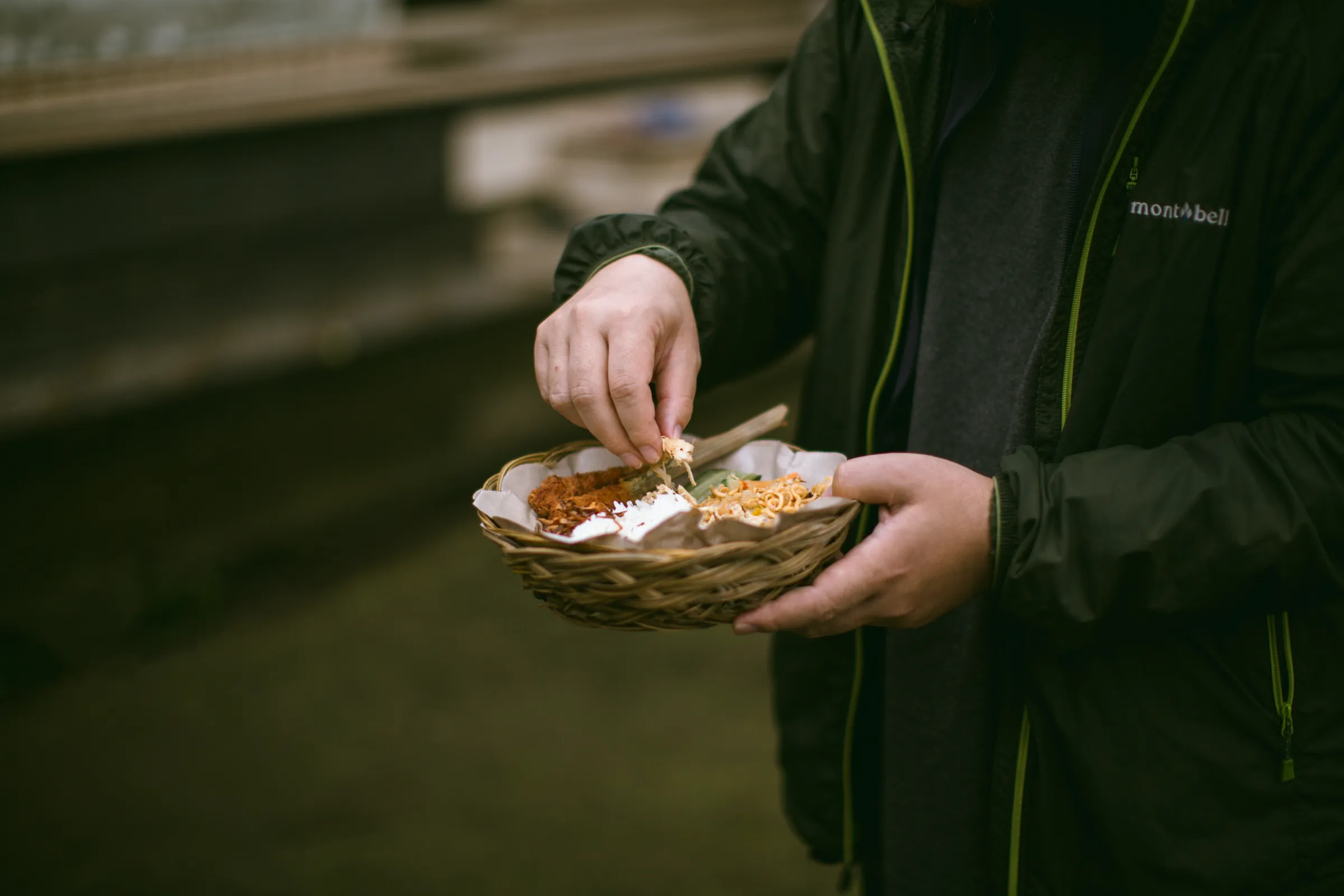 A guest eating by hand from a woven basket of rice, noodles and sate